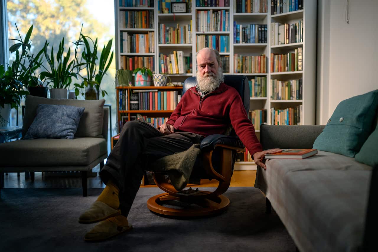 A man wearing a red jumper with a white beard sits on a chair inside his home. There is a colourful bookcase behind him and potted plants.