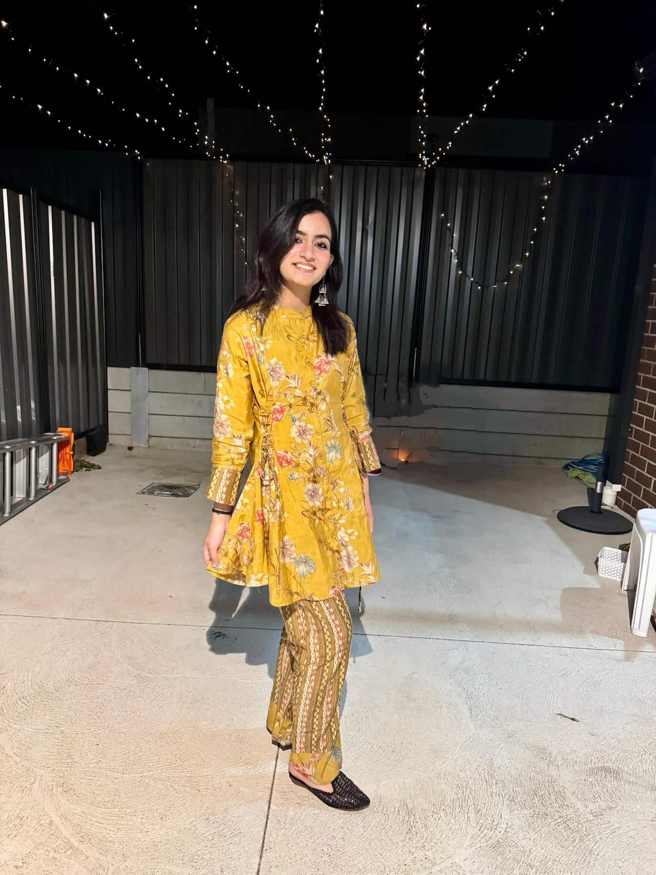 A young woman in a traditional Indian yellow suit poses for the camera as she stands in a backyard decorated with fairy lights.