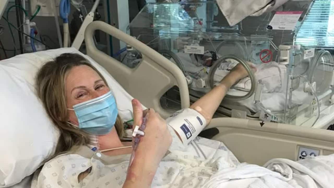 a woman in hospital bed with a mask on, putting her arm into an incubator with babies in it