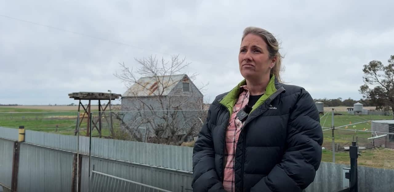 A woman in a puffer jacket stands on a deck in front of a paddock, her face expressing sadness.