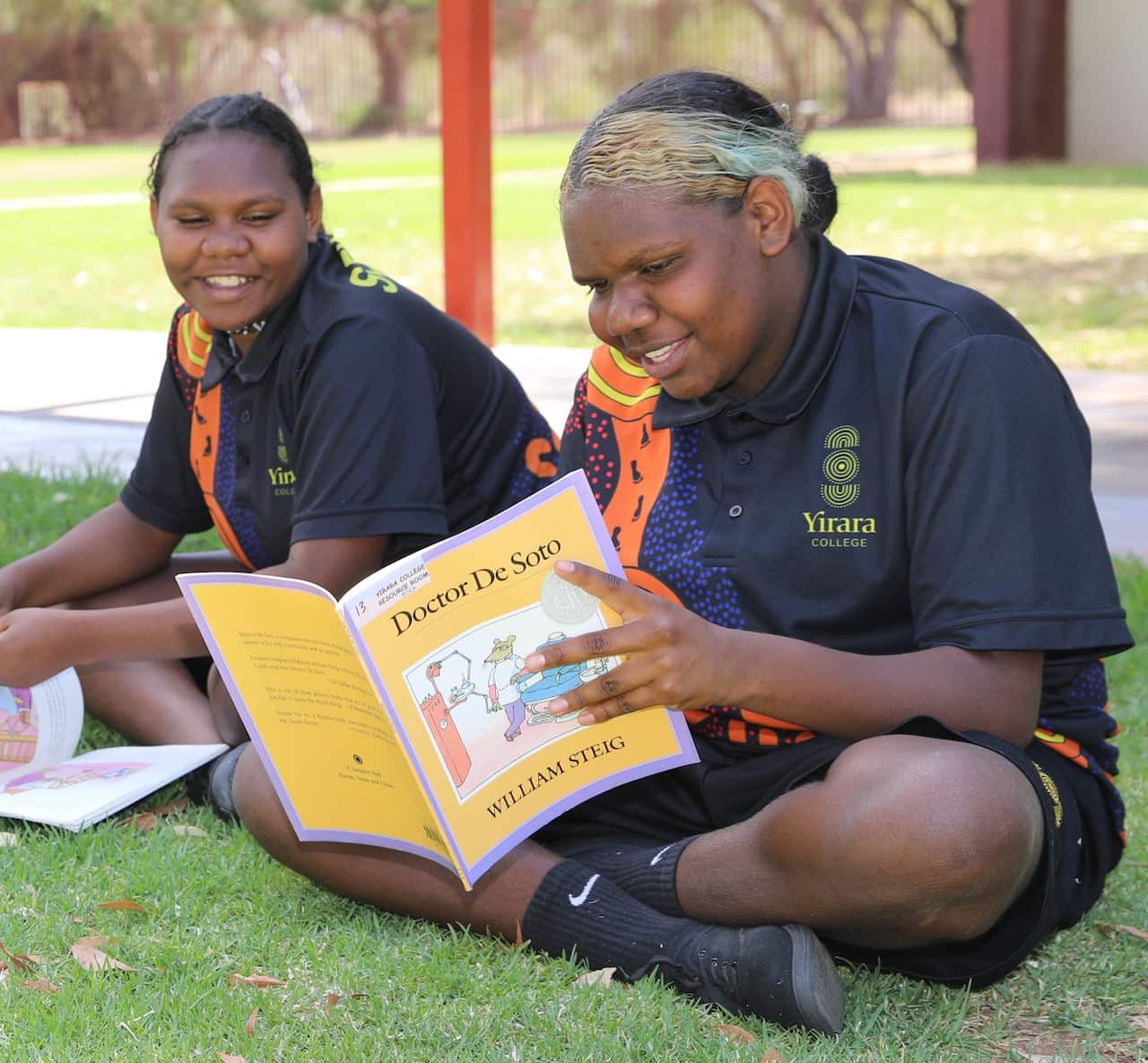 Two female students in school uniform sitting cross legged outside on grass reading books.