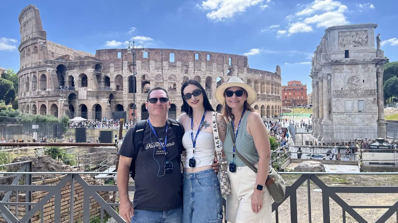 a middle aged man, young woman and middle aged woman stand arm in arm in front of the Colosseum in Rome