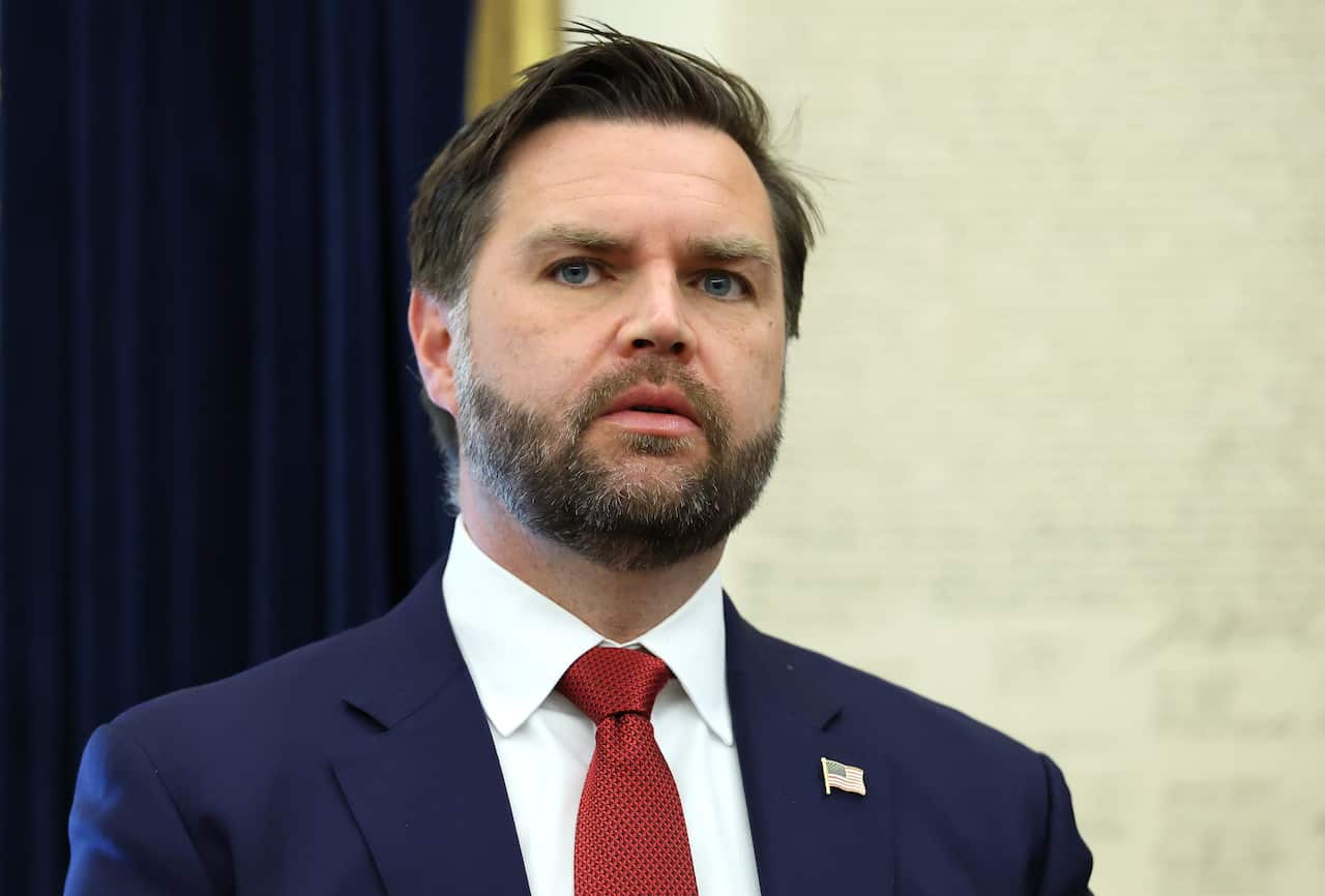 JD Vance, wearing a blue blazer, white shirt, and red tie. The blazer has an American flag pin on it.