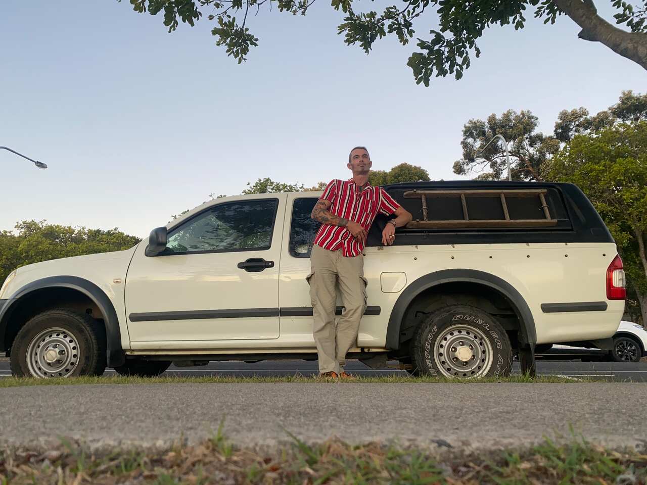 A man in a red shirt with white stripes leans on the side of his white ute.