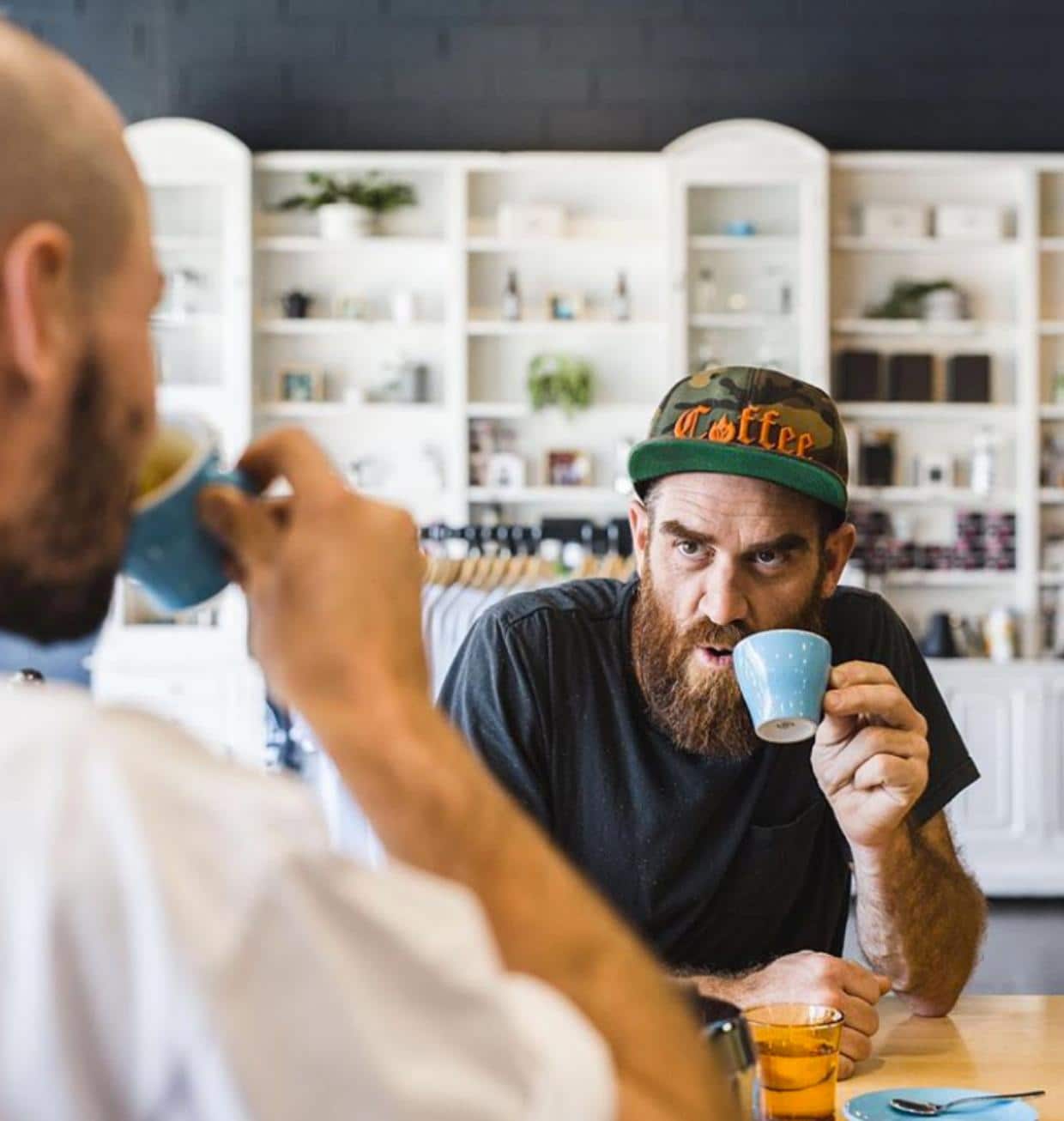 A man in a t-shirt and cap drinking a coffee from a small blue cup. 