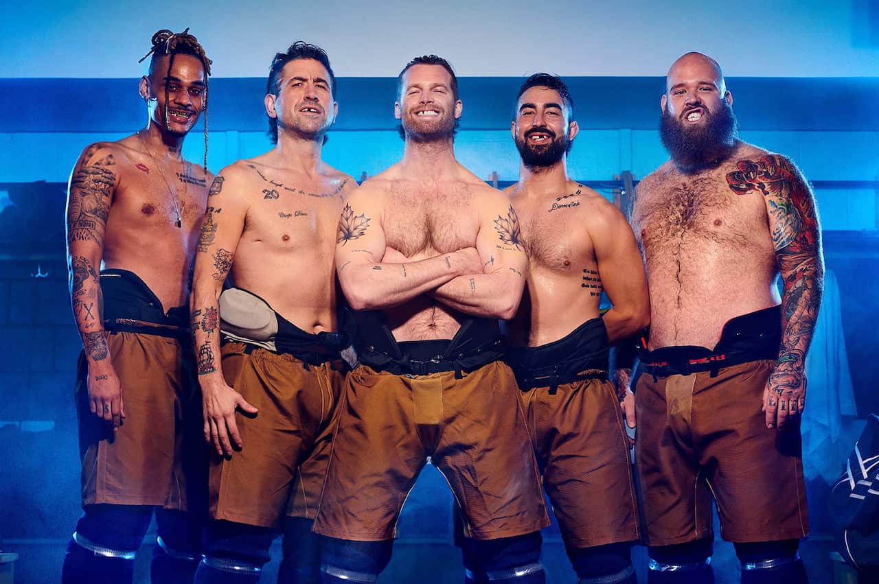Five men with bare chests and wide grins stand in a locker room. 
