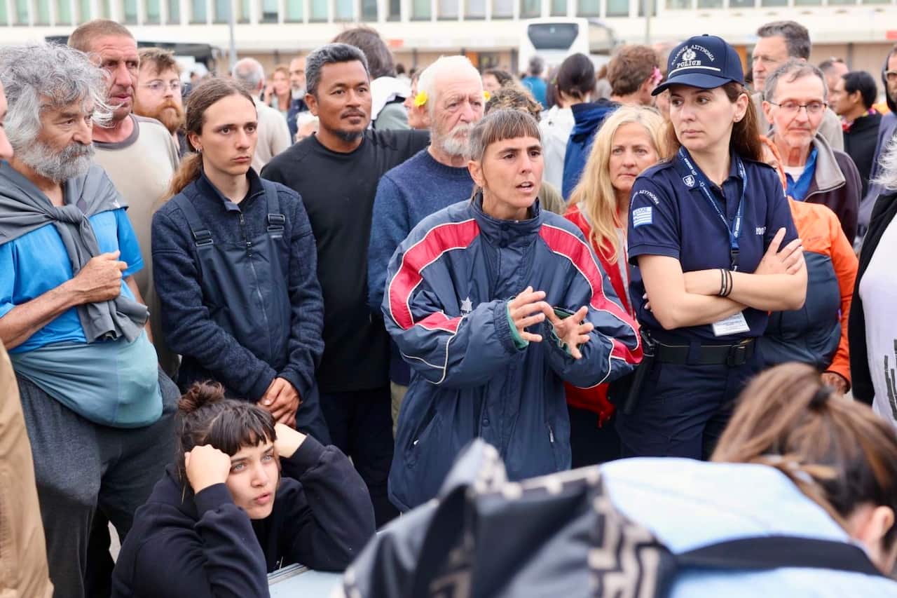 A crowd of people, including police officers, gather in a crowd at a port. Two women hold their hands up.