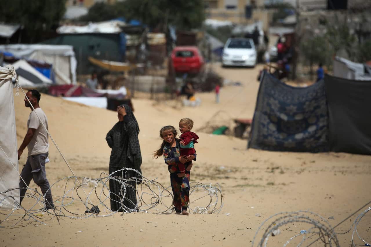 Small children stand next to barbed wire with tents and vehicles in the background. 