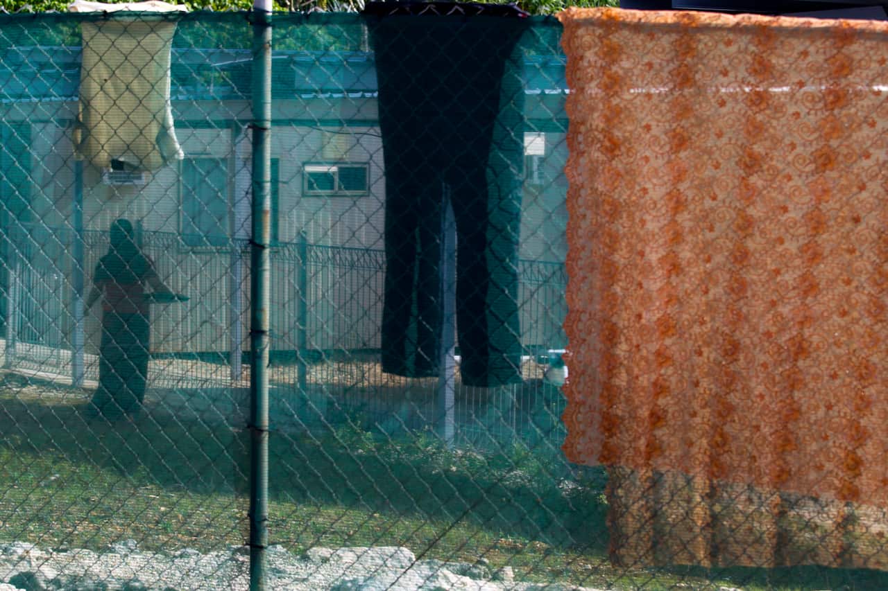 The silhouette of a woman carrying a washing basket behind a wire fence.