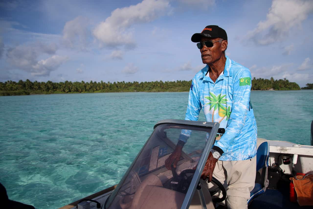 An elderly man wearing a cap, sunglasses, and a light blue tropical shirt steers a boat across clear turquoise water toward a lush, palm-lined island shore.