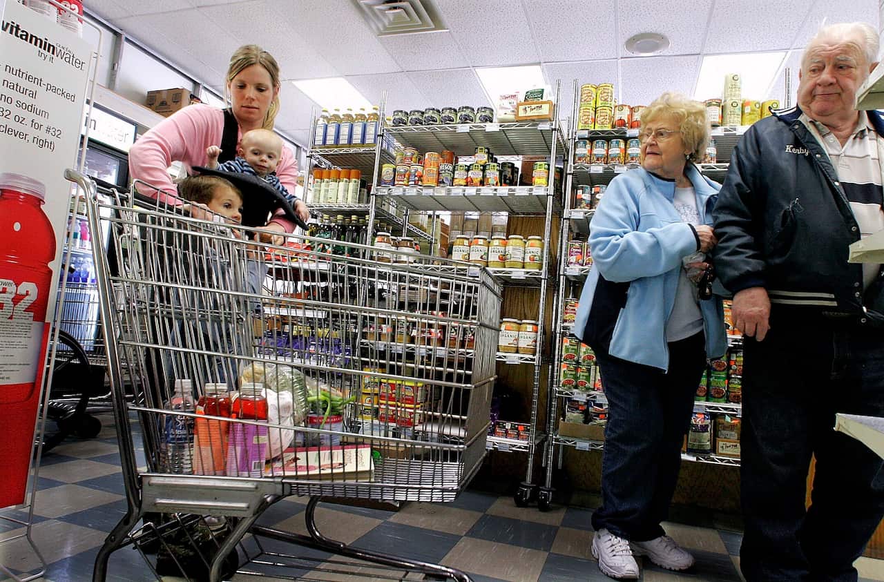 A woman navigates a grocery store with a trolley, while carrying a baby and walking with a young child, as an older couple look on.