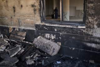 Two people pray inside a burned building. Outside the building, a burned stone with an Arabic inscription is seen.