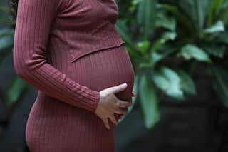 A woman in a knitted purple dress holds her pregnant belly in front of a vine trellis