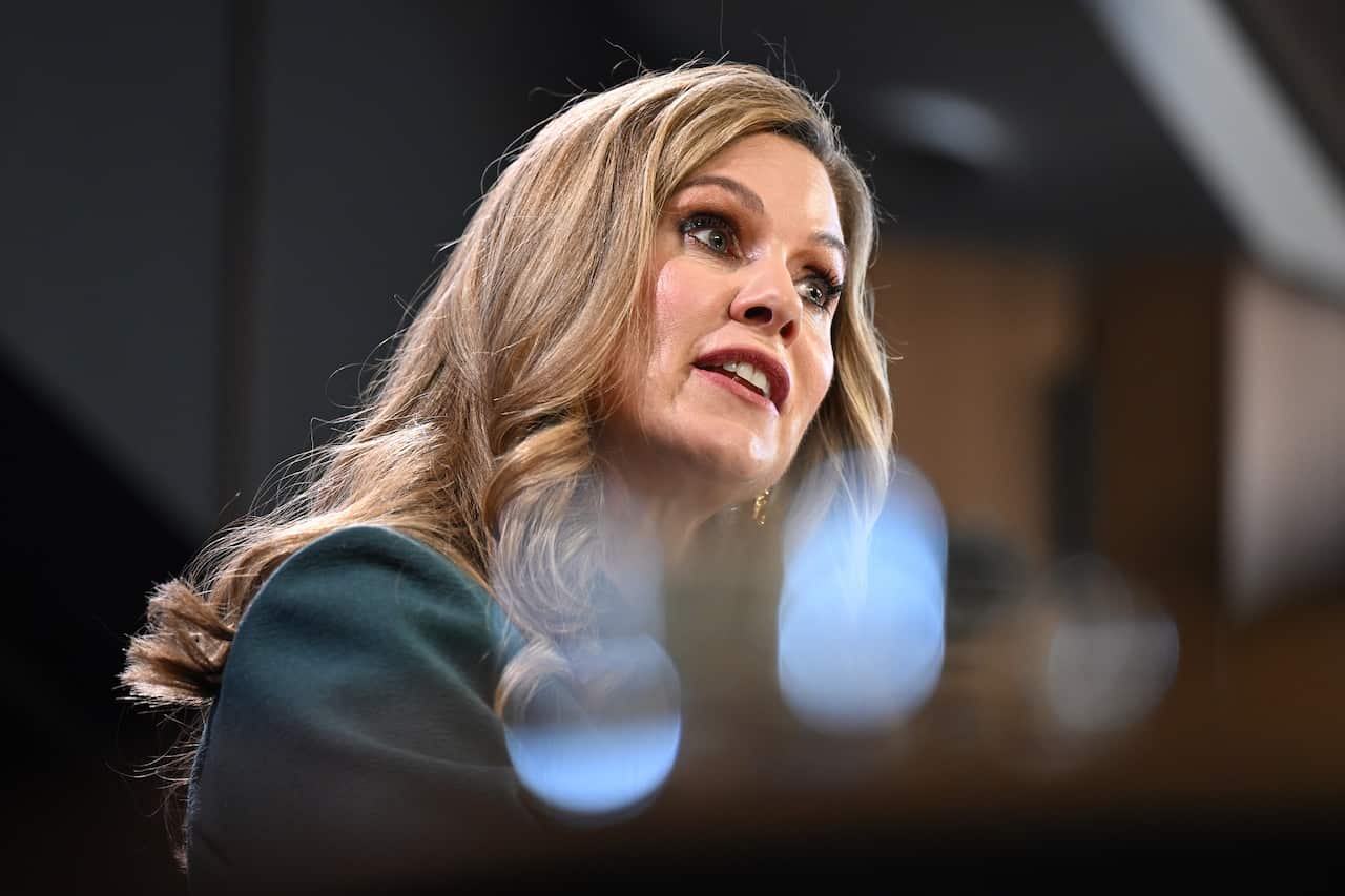 A woman with blonde hair and a dark green jacket looks to her right while speaking.