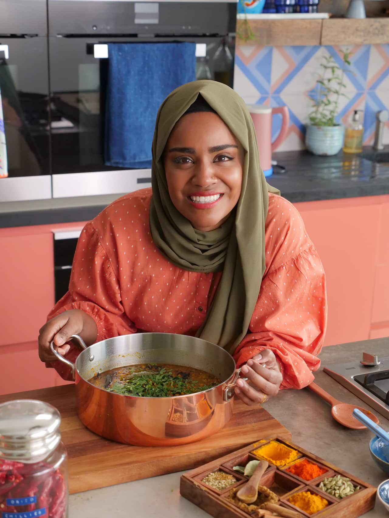 A woman in an orange top and green headscarf stands behind a kitchen bench, holding a copper pot of dhal. A box of various spices sits on the bench. 