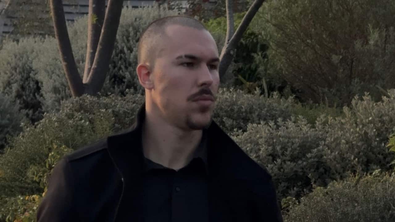 a young man with a shaved head, eyebrow slit and facial hair in a black coat stands in front of shrubs