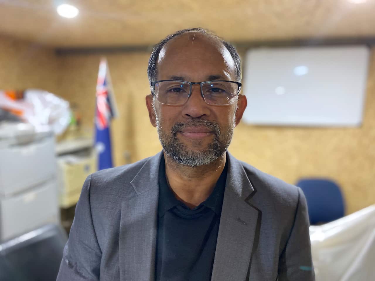 A man wearing glasses, a dark shirt and grey jacket inside a room