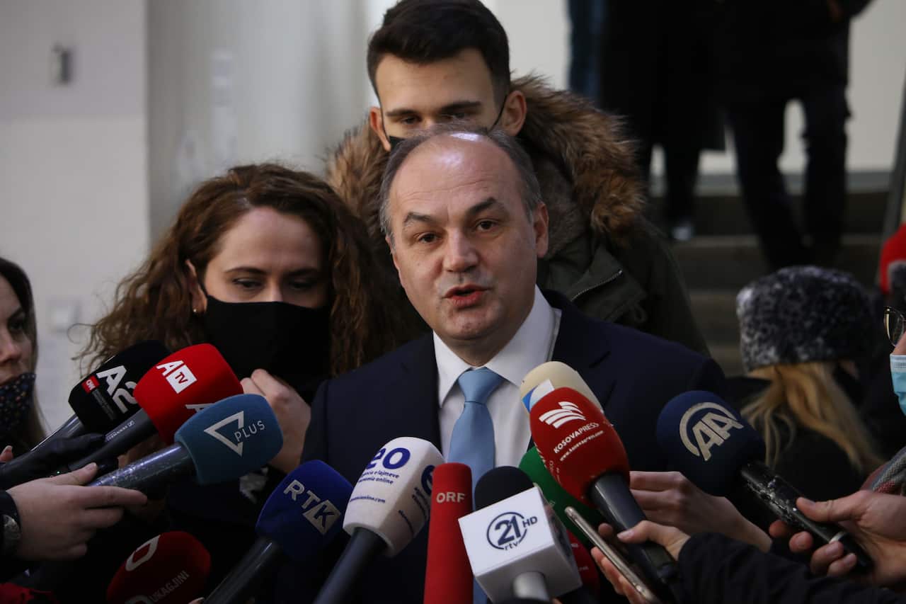 A man in a suit and light blue tie speaks into a cluster of microphones held by journalists during a media briefing in a crowded indoor setting.