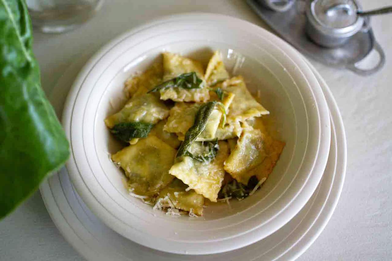 A low white bowl sits on a matching white plate. The bowl holds filled pasta squares, with sage leaves and cheese on top. 