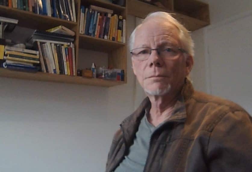 A middle aged man in a jacket in a home office with a bookshelf behind him.