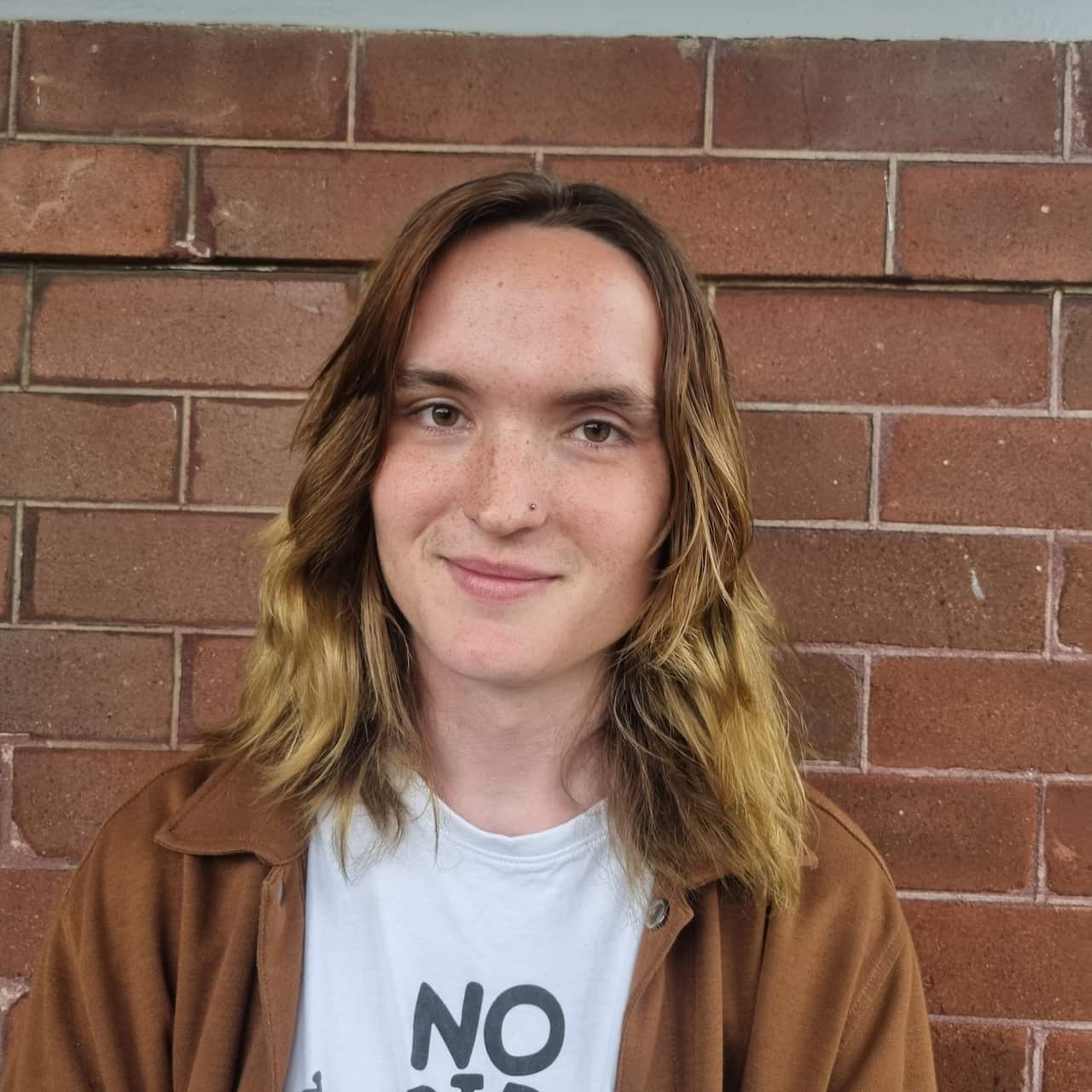 A woman with shoulder-length blonde hair in a brown shirt smiles while standing in front of a brick wall