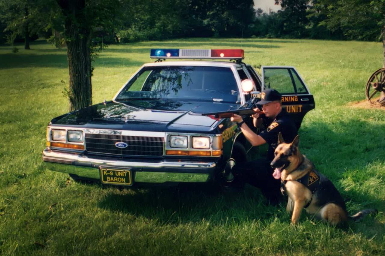 A man dressed as a US sheriff kneels down next to a patrol car. A German Shepherd dog sits beside him.