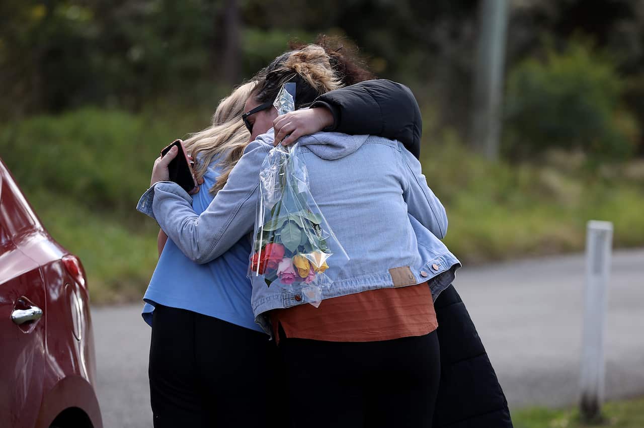 A group of women embrace one another, carrying flowers. 