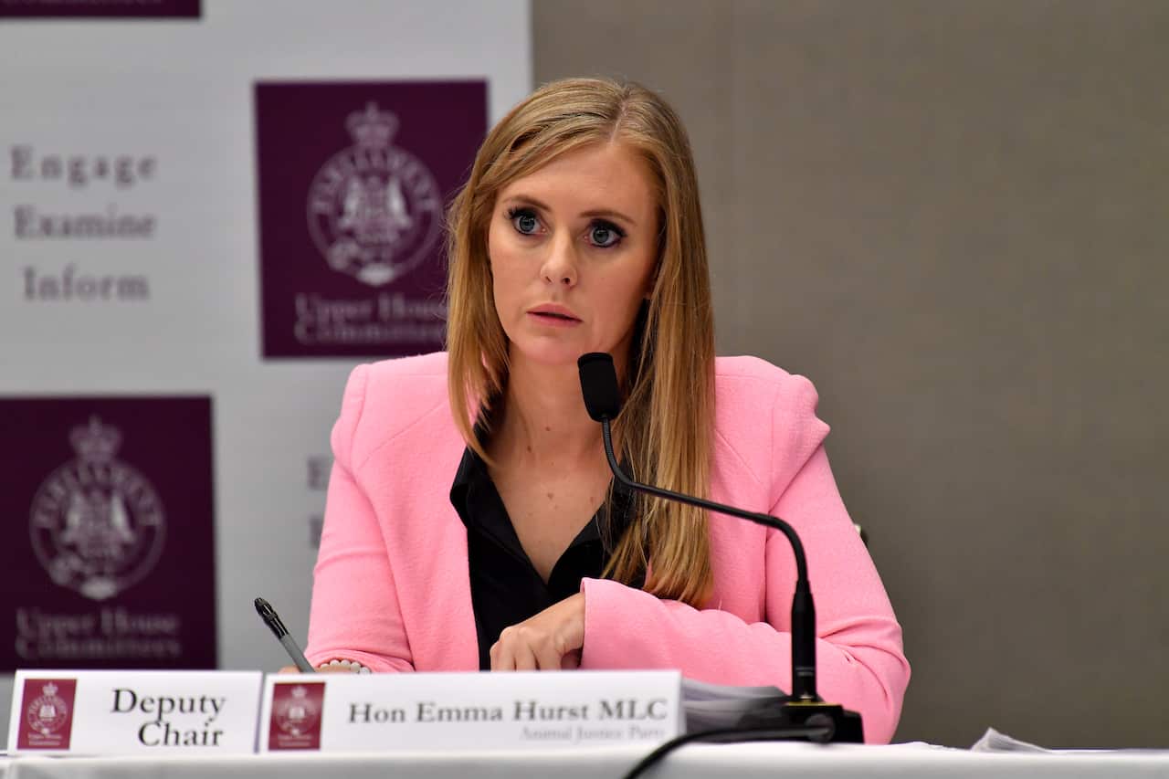 A blonde woman in a bright pink blazer and black top is seated at a table with a microphone, looking intently to the side. Behind her, a purple banner displays the logo and name "Upper House Committees."