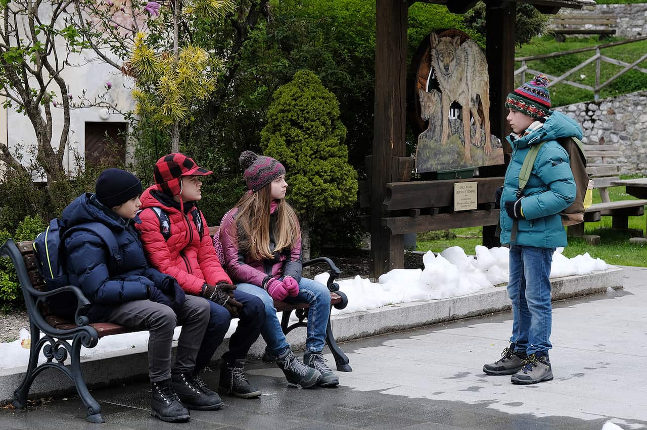 Three children in winter clothing sit on a bench. A fouth child stands in front of them, and appears to be talking with one of the children on the bench.