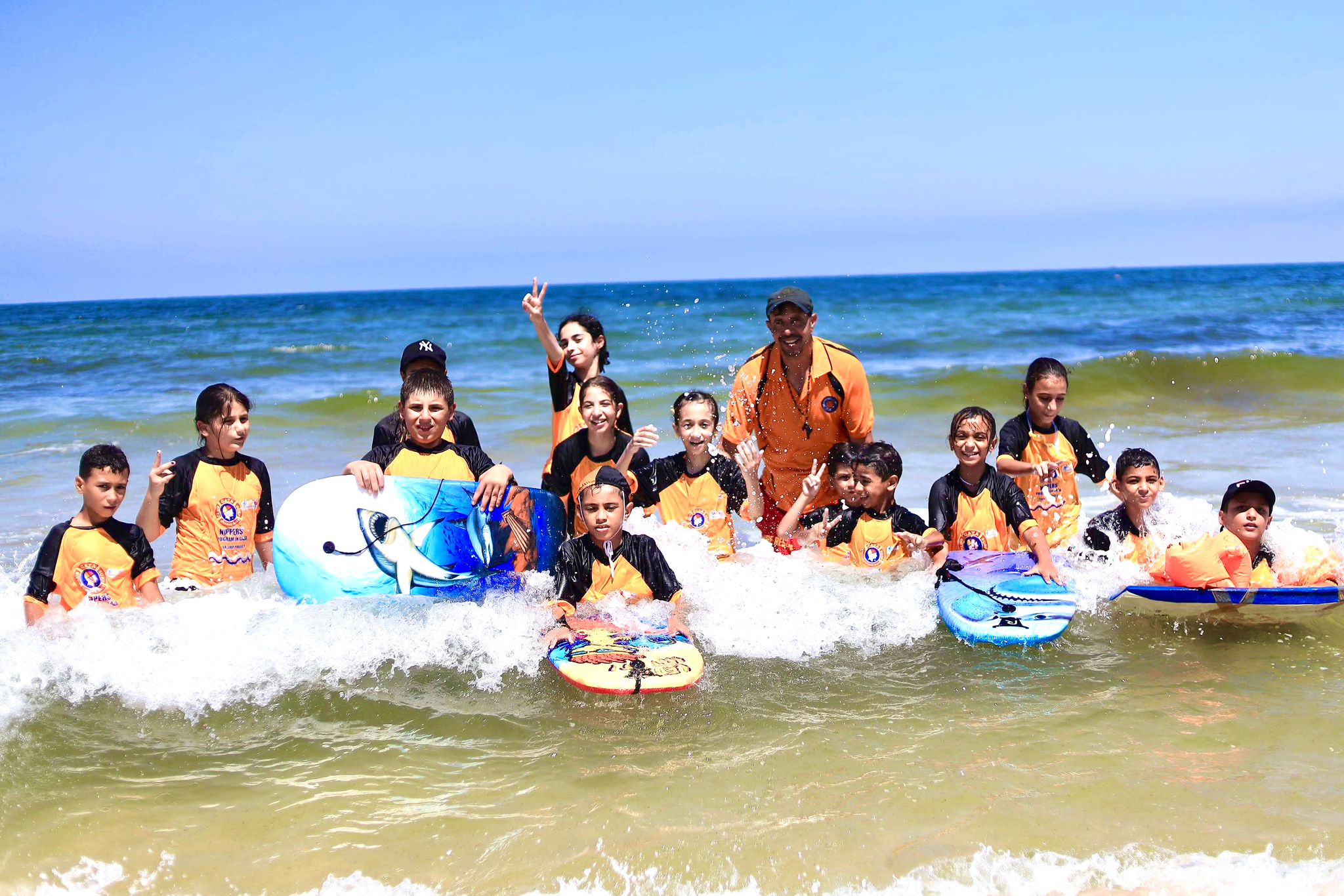 A joyful group of children and an adult instructor in the ocean waves on a sunny day, with the children wearing matching black and orange surf tops.