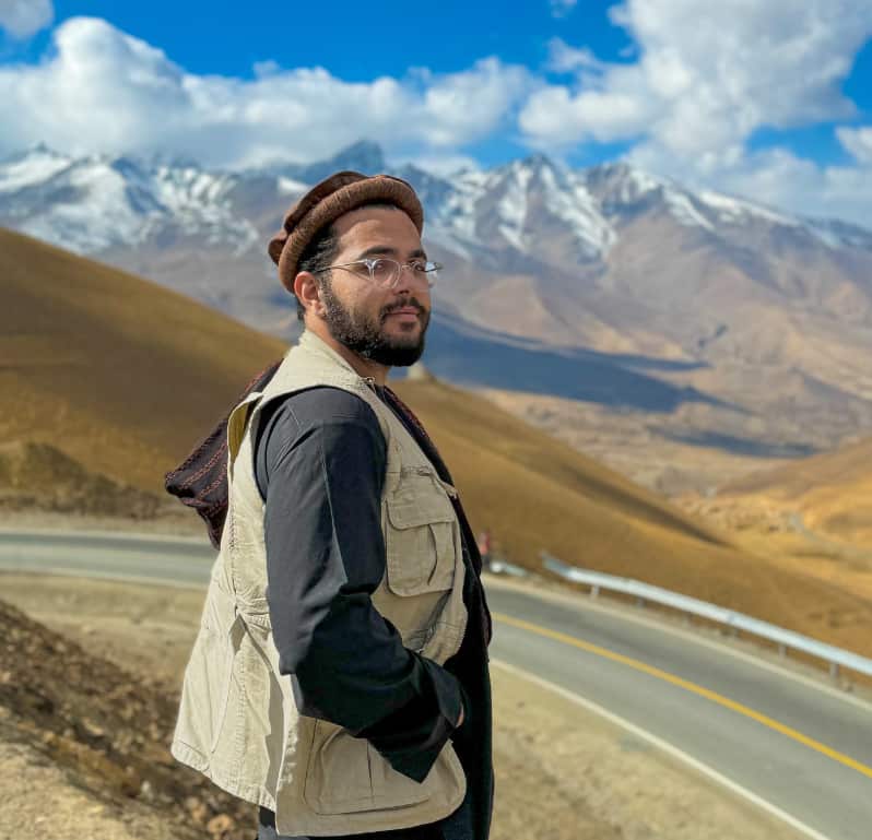 A man dressed in a vest and a traditional Afghan hat stands by the roadside with sandy hills and snow-capped mountains in the distance.