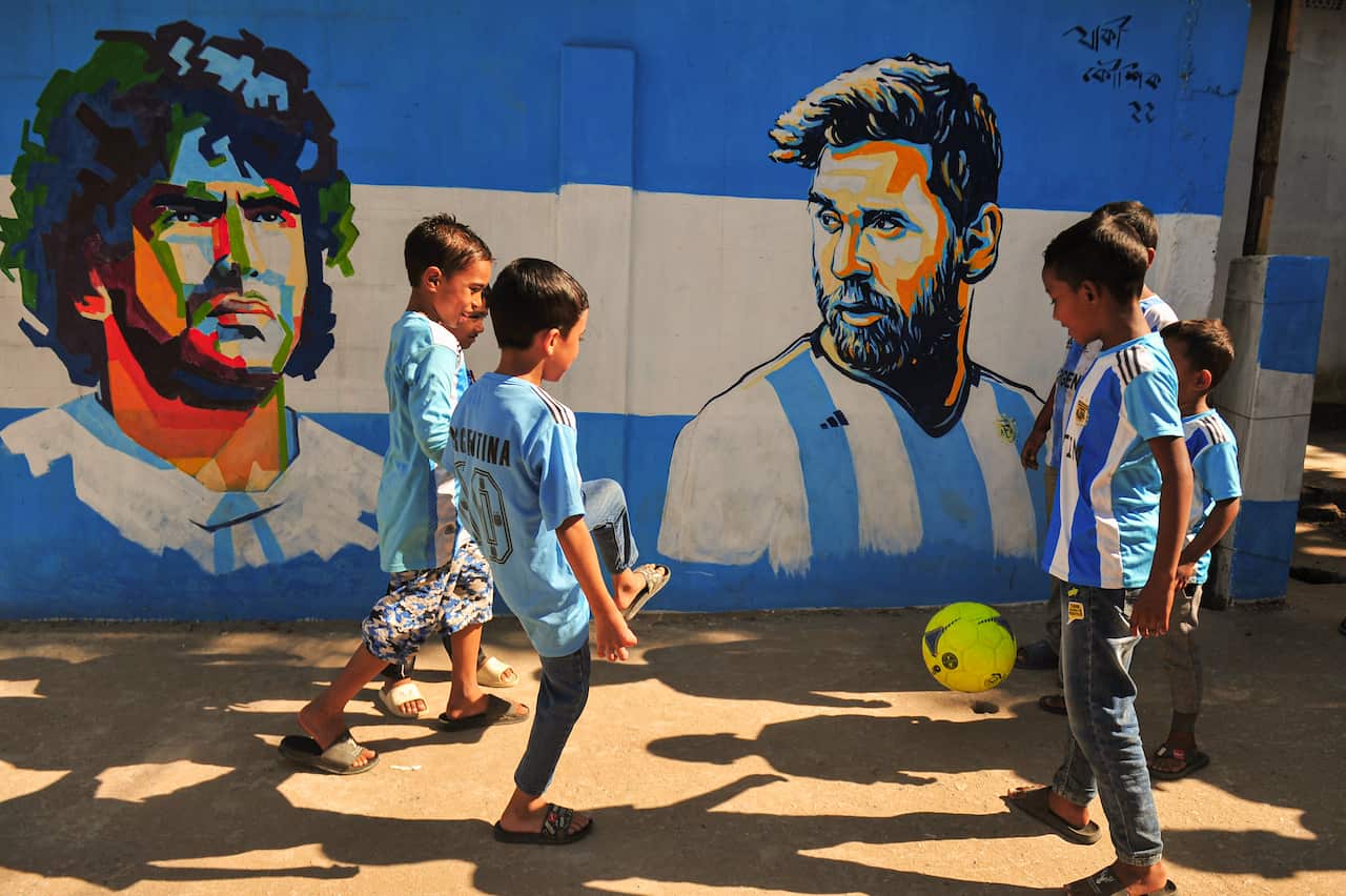 Children Fans of Argentina play football on the streets in front of a blue and white mural.