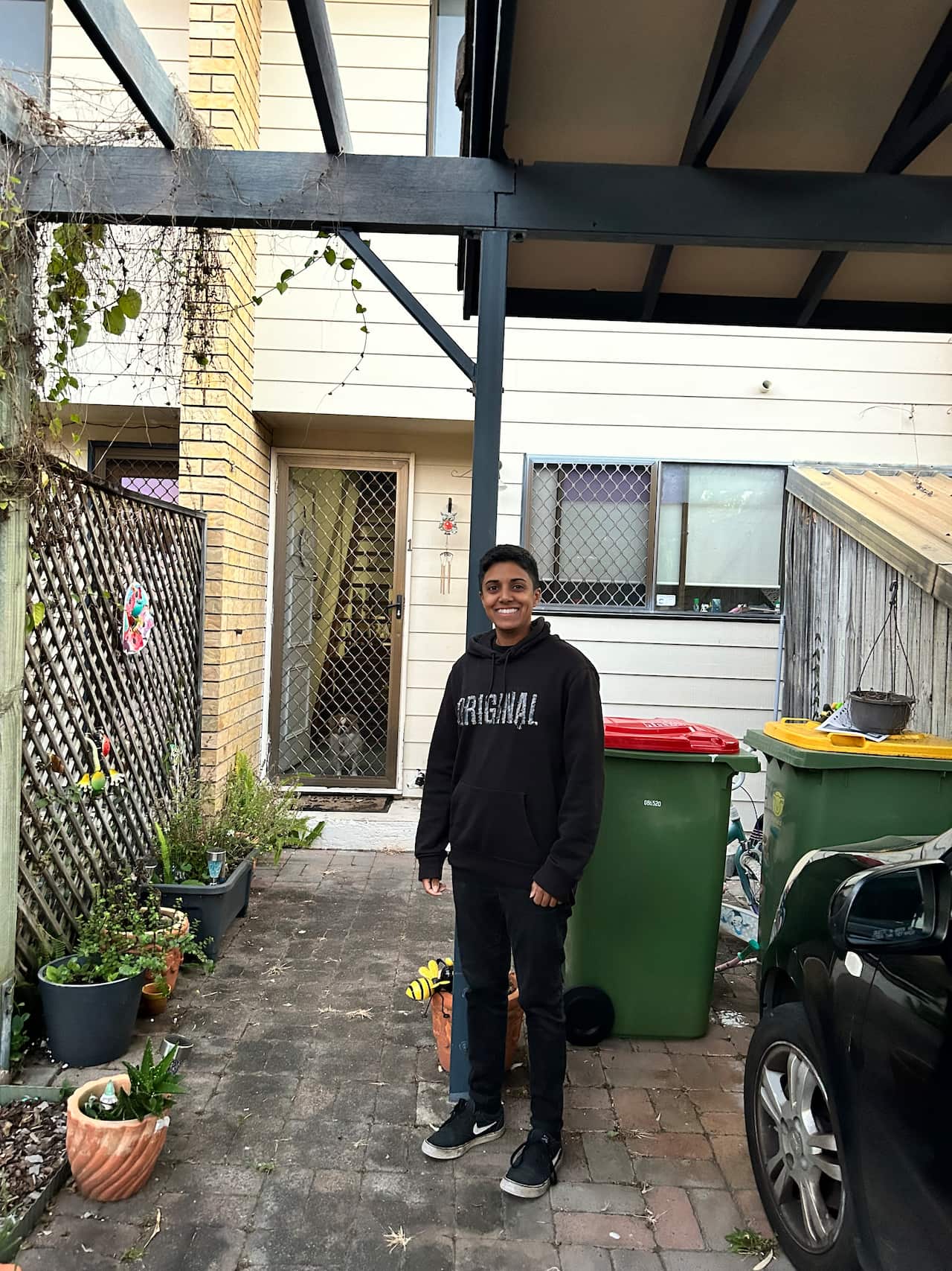 A woman wearing a black jumper and jeans stands in a small courtyard outside a home.