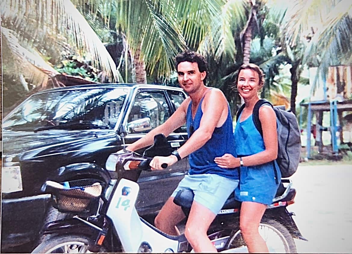 A man and a woman wearing blue clothes smile as they sit on a motorbike in a tropical setting.