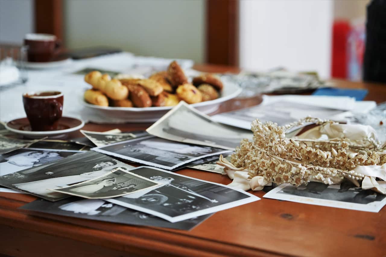 On a wooden table, dozens of black-and-white photos are strewn around, showing glimpses of weddings; in the background, a cup of coffee and Greek sweets sit on a plate. In the foreground, the flower crowns known as a stefana in Greek sits atop the photos. 
