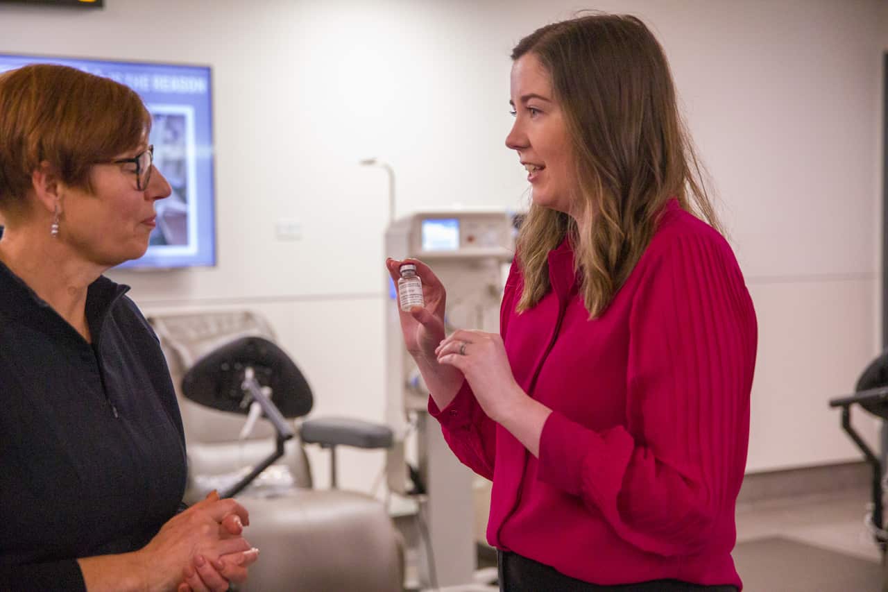 A woman in a dark pink blouse, holding up a vial of plasma as she speaks to another woman.