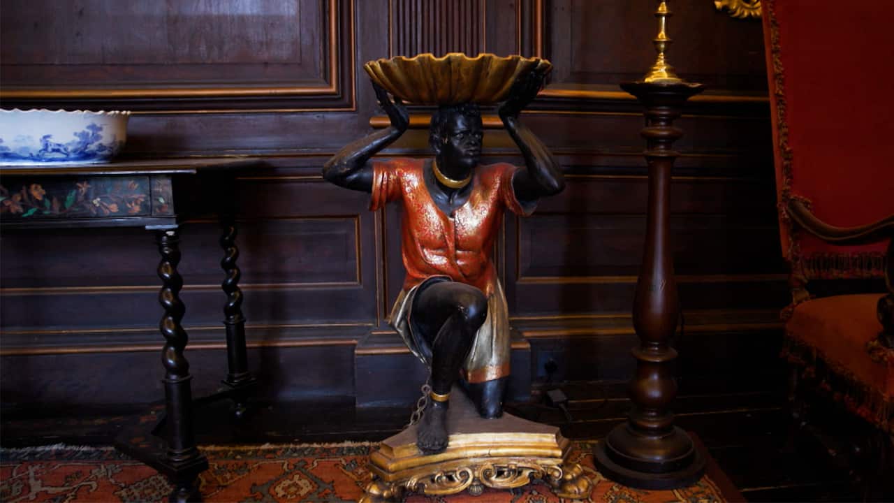 A carved statue of a Black person chained at the ankles and neck, holding a scallop shell on its head is displayed in a room with wooden walls. 