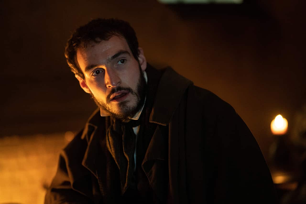 A man in 19th century clothing is seen, head and shoulders, in a dark room. One side of his face is in the shadows as he looks to one side. 