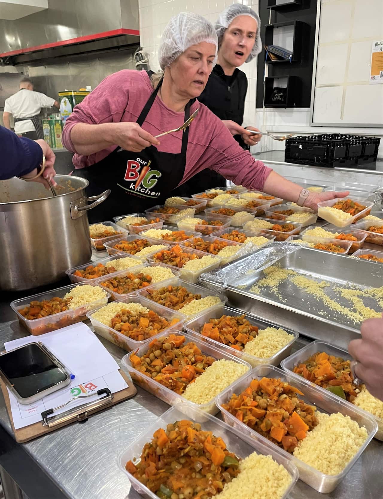 Two women in hair nets ladle food into plastic meal boxes on a stainless steel table.