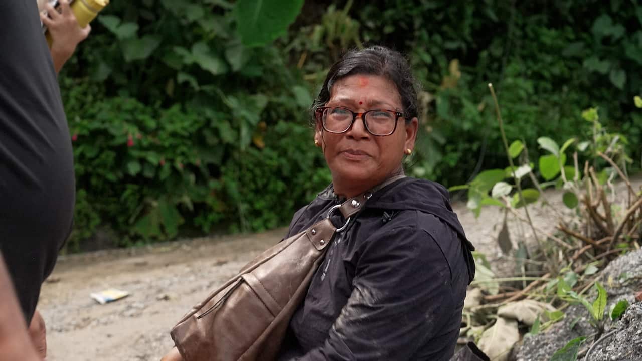 An older woman of South Asian descent wearing glasses, a black jacket and a bag. She's leaning against a rock outside.