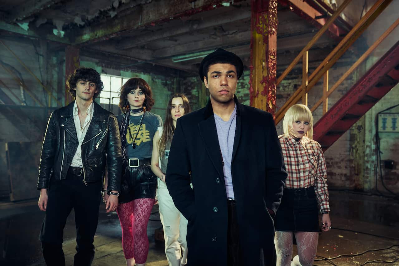 Three women and two men stand in a derelict warehouse wearing 80s new wave style outfits