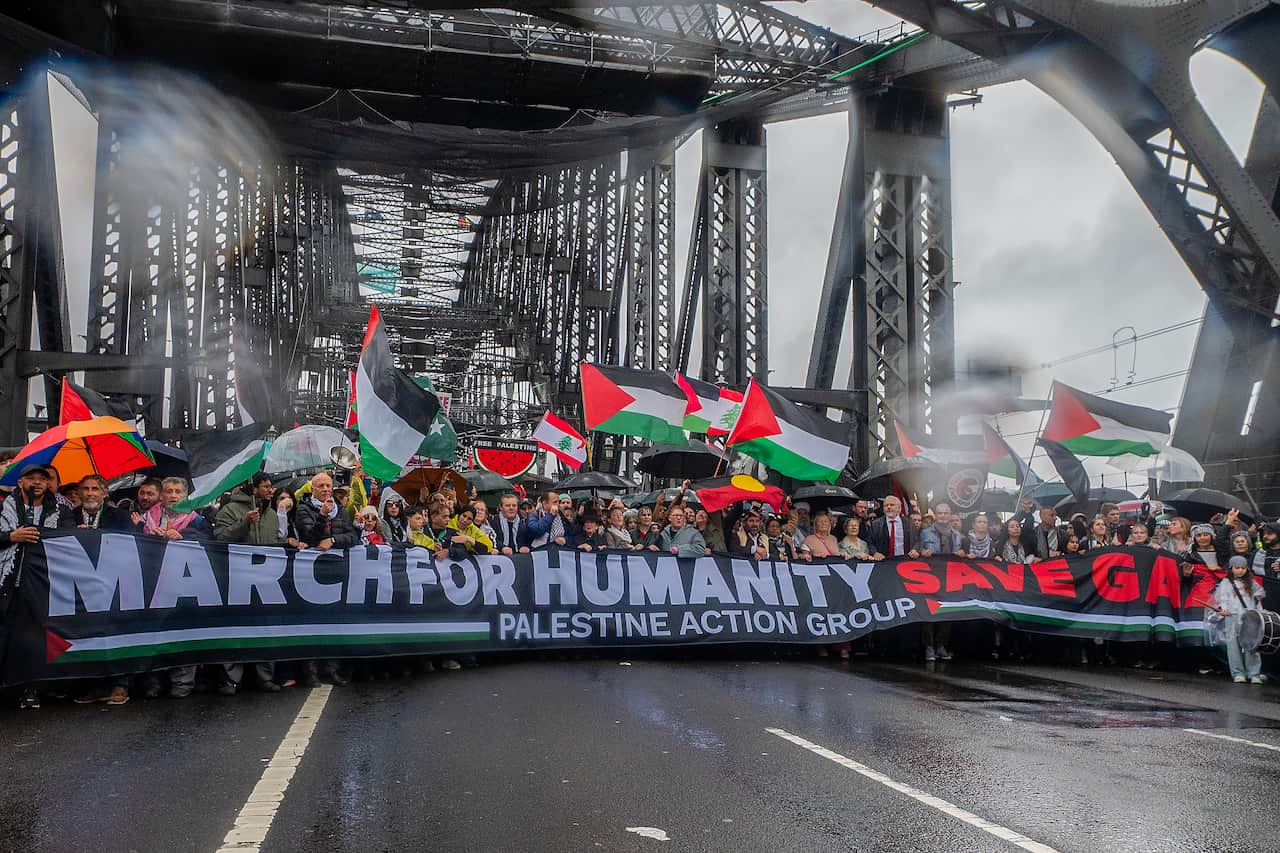 A large group of people marching across the Sydney Harbour Bridge, many holding Palestinian flags.