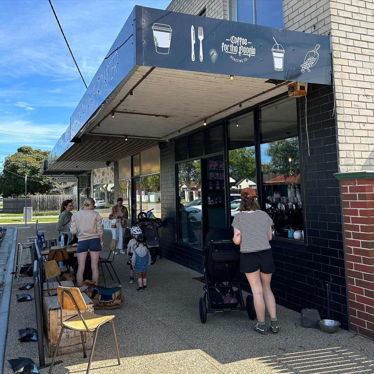The outside of a cafe, where women with prams and young children meander near tables and chairs.