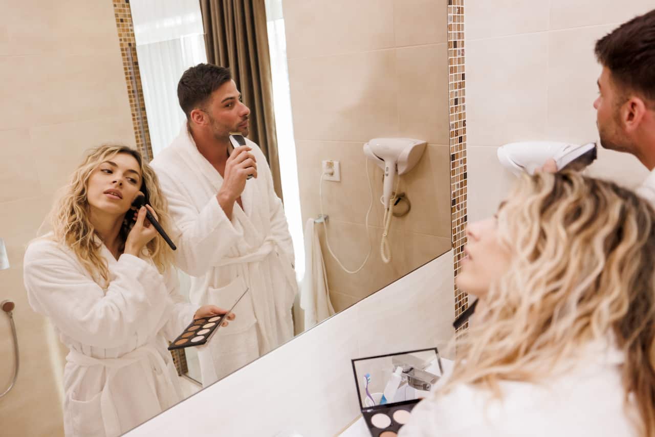 Man and woman in bathroom, looking in mirror as woman applies makeup and man shaves.