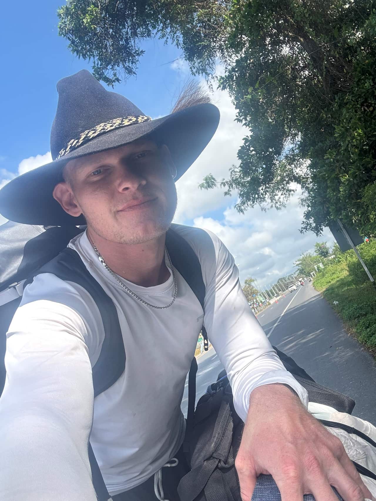 A man in a long-sleeved white t-shirt and wearing a cowboy hat poses for a selfie on the side of a road.