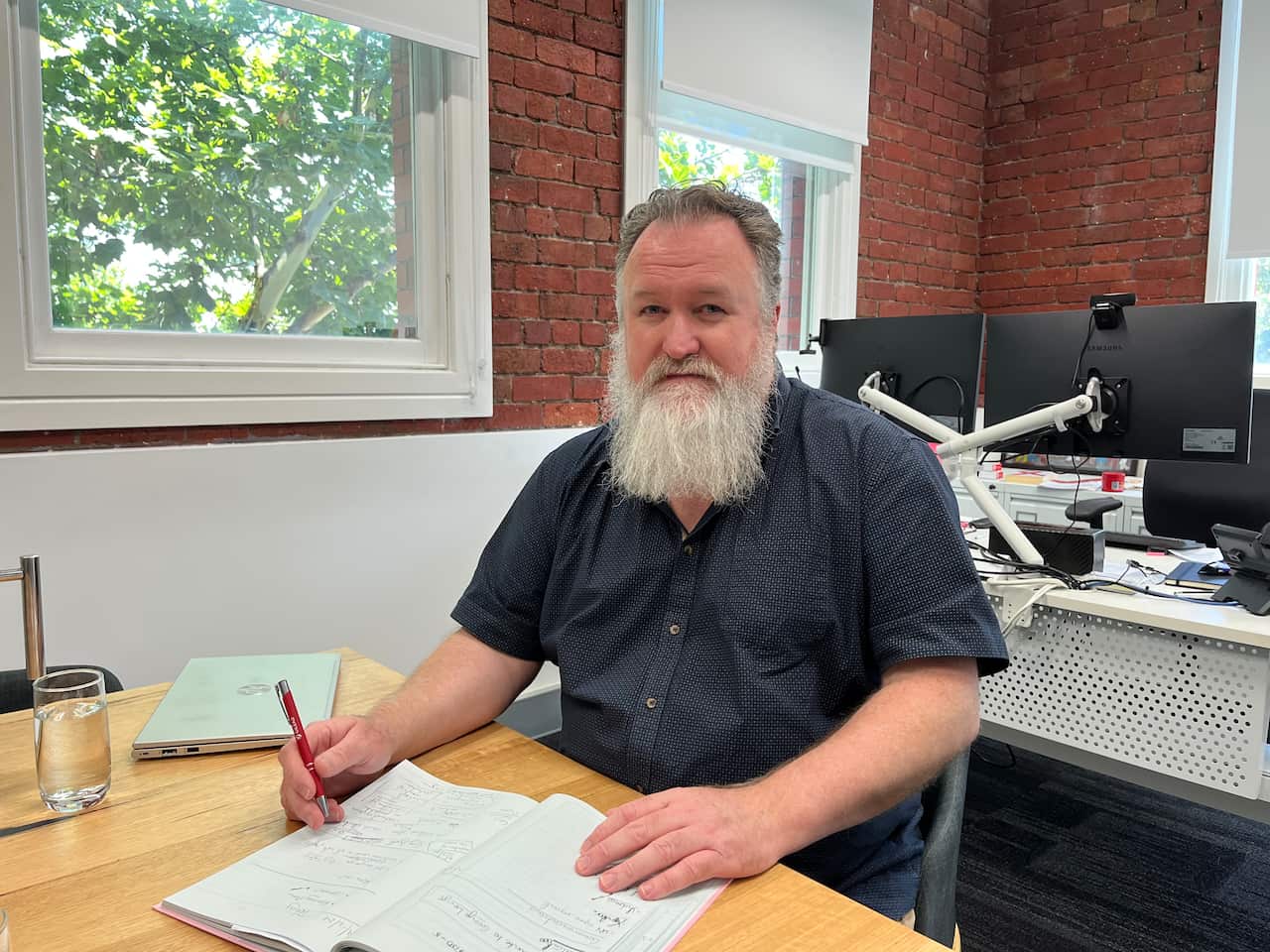 A man in a black shirt with a long beard is sitting at his desk.