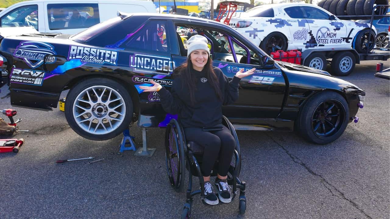 a woman in black clothes and a grey beanie sitting in a wheelchair smiles in front of a drifting car in a caryard