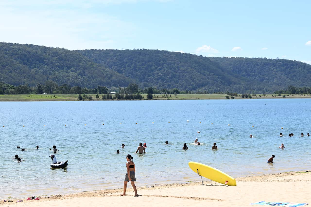 People swimming in a wide river. A yellow surfboard is on the sand in the foreground and in the distance there are green fields and forest.