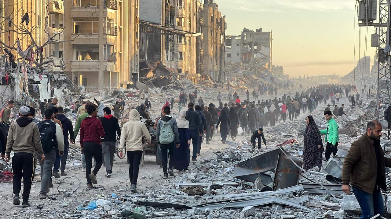 A line of hundreds of people walk through the rubble of destroyed buildings.