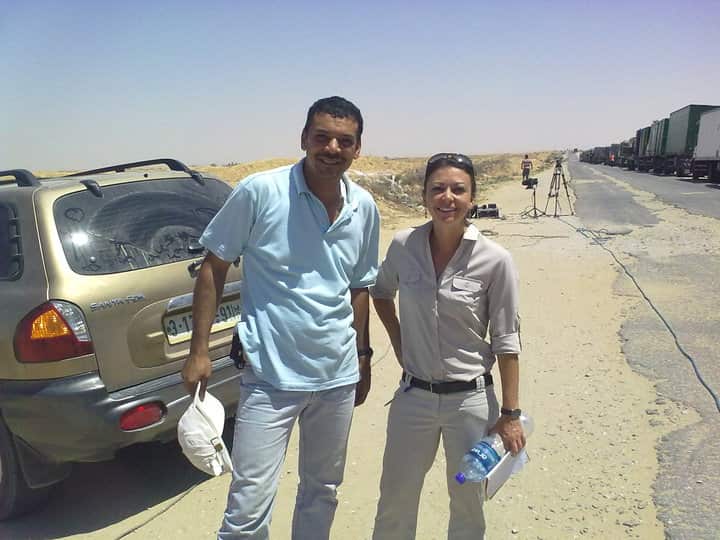 A woman in a cream-coloured shirt (right) stands next to a man in a light-blue shirt next to a road. There is a four-wheel-drive vehicle parked next to them.